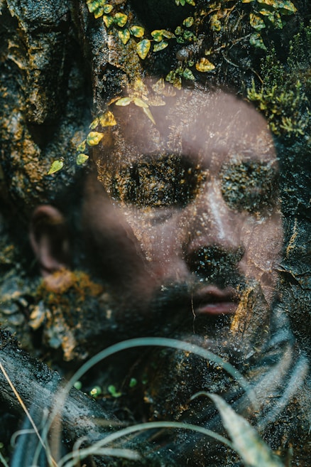 A surreal blend of a human face with a textured, earthy surface, interwoven with scattered leaves and greenery. This image creates an ethereal and dreamlike composition.