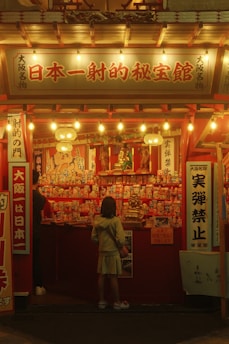 A child stands in front of a brightly lit booth, adorned with various traditional Japanese decorations and an assortment of small, colorful items on display. The booth has Japanese signage with illuminated lanterns hanging from the roof.