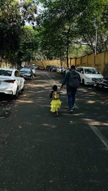 A child wearing a bright yellow dress and a shiny backpack is walking hand in hand with an adult down a tree-lined street. The road is flanked by parked cars on both sides, and large trees provide ample shade. The scene exudes a sense of everyday life, with autumn leaves scattered on the pavement.