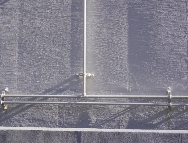 A textured gray wall with a vertical and two horizontal white pipes arranged in a T-shape pattern, casting shadows on the wall. The pipes are attached to the wall with clamps.