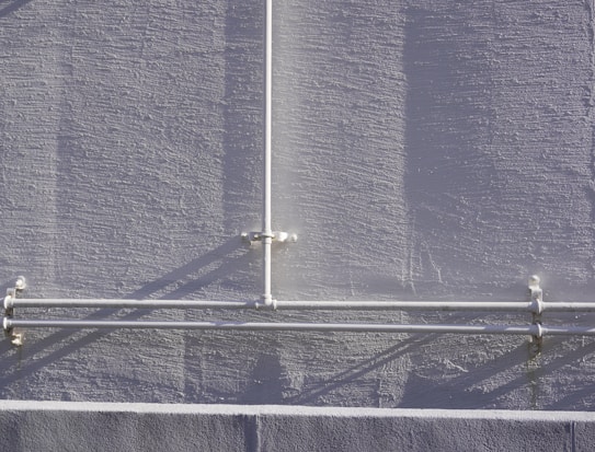 A textured gray wall with a vertical and two horizontal white pipes arranged in a T-shape pattern, casting shadows on the wall. The pipes are attached to the wall with clamps.