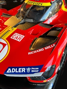A red racing car prominently featuring the Ferrari logo on the windshield area. The car has decals and sponsorship logos, such as Richard Mille and Adler Pelzer Group, and is labeled with 'Hyper Car' on the side. The sleek aerodynamic design and the bright color scheme give it a dynamic and high-performance appearance.