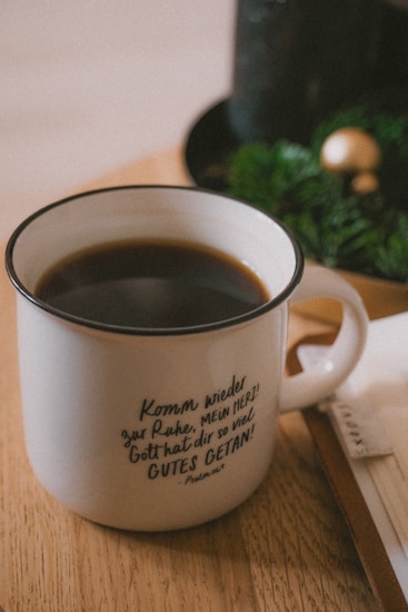 A white ceramic mug with a German inscription on it sits on a wooden table. The mug is filled with black coffee. In the background, there is a hint of green foliage and a golden spherical decoration, suggesting a cozy and festive atmosphere.