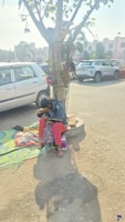 A person is sitting on a mat by a tree in a parking lot with cars in the background. They appear to be holding a child. The tree has a fence around its base and there are items like a bottle near them. The ground is paved, and the area is well-lit, suggesting daytime.