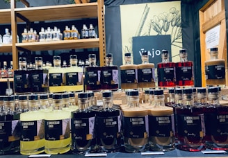 Bottles of various flavored liqueurs are displayed in rows, each labeled with distinct flavors such as Limoncello, Haselnuss, and Peche Rouge. The backdrop includes a shelf filled with additional bottles of spirits and a decorative sign featuring botanical illustrations.