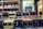Bottles of various flavored liqueurs are displayed in rows, each labeled with distinct flavors such as Limoncello, Haselnuss, and Peche Rouge. The backdrop includes a shelf filled with additional bottles of spirits and a decorative sign featuring botanical illustrations.