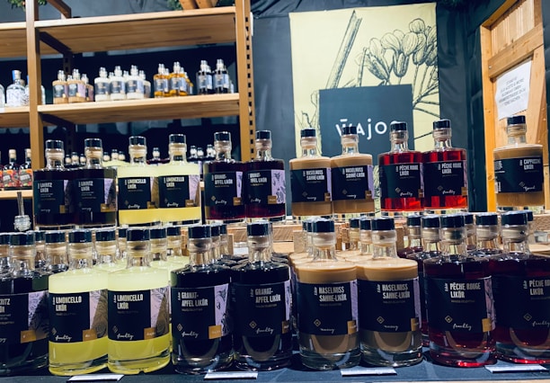 Bottles of various flavored liqueurs are displayed in rows, each labeled with distinct flavors such as Limoncello, Haselnuss, and Peche Rouge. The backdrop includes a shelf filled with additional bottles of spirits and a decorative sign featuring botanical illustrations.