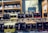 Bottles of various flavored liqueurs are displayed in rows, each labeled with distinct flavors such as Limoncello, Haselnuss, and Peche Rouge. The backdrop includes a shelf filled with additional bottles of spirits and a decorative sign featuring botanical illustrations.