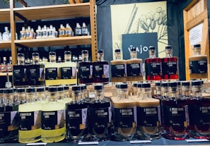 Bottles of various flavored liqueurs are displayed in rows, each labeled with distinct flavors such as Limoncello, Haselnuss, and Peche Rouge. The backdrop includes a shelf filled with additional bottles of spirits and a decorative sign featuring botanical illustrations.