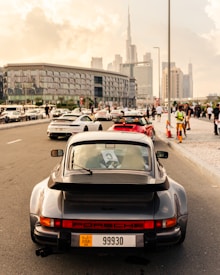 A classic Porsche car with a Dubai license plate is parked on a bustling city street. Numerous luxury sports cars are lined up along the road, with people walking nearby. The skyline in the background features tall buildings, including a prominent skyscraper under a partly cloudy sky.