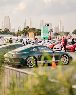 A green sports car is parked among a collection of luxury vehicles, with a scenic urban skyline of tall skyscrapers and palm trees in the background. The foreground includes green foliage, a traffic cone, and multiple people walking around.