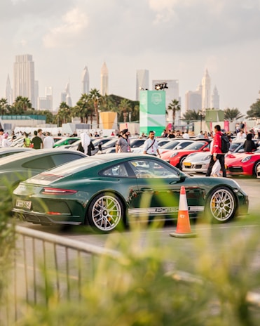 A green sports car is parked among a collection of luxury vehicles, with a scenic urban skyline of tall skyscrapers and palm trees in the background. The foreground includes green foliage, a traffic cone, and multiple people walking around.