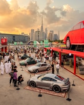 A vibrant gathering at an outdoor car exhibition featuring luxury sports cars on display, surrounded by a large crowd of people. The event is set against the backdrop of a modern city skyline with a prominent skyscraper. The setting sun casts a warm glow over the scene, adding to the lively atmosphere. A large red car-shaped structure is elevated on a platform above the crowd.