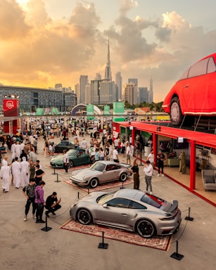 A vibrant gathering at an outdoor car exhibition featuring luxury sports cars on display, surrounded by a large crowd of people. The event is set against the backdrop of a modern city skyline with a prominent skyscraper. The setting sun casts a warm glow over the scene, adding to the lively atmosphere. A large red car-shaped structure is elevated on a platform above the crowd.