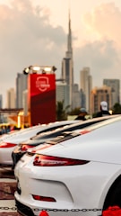 A lineup of sleek, high-performance sports cars parked with a modern city skyline in the background. The cars are predominantly white and red, with a luxury brand logo visible. The sky is slightly overcast, providing a dramatic backdrop to the scene.