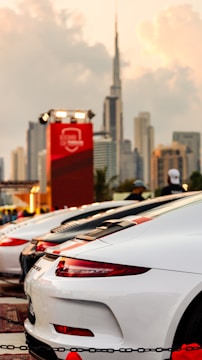 A lineup of sleek, high-performance sports cars parked with a modern city skyline in the background. The cars are predominantly white and red, with a luxury brand logo visible. The sky is slightly overcast, providing a dramatic backdrop to the scene.