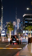 An evening cityscape featuring a vibrant street with a mix of modern skyscrapers, palm trees, and people dressed in traditional attire. A black SUV is traveling down the street, which is lined with illuminated buildings and decorative lights. The scene includes a prominent, illuminated tower in the background.