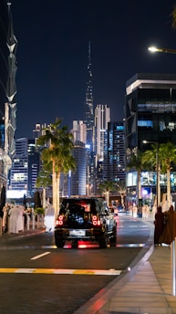 An evening cityscape featuring a vibrant street with a mix of modern skyscrapers, palm trees, and people dressed in traditional attire. A black SUV is traveling down the street, which is lined with illuminated buildings and decorative lights. The scene includes a prominent, illuminated tower in the background.
