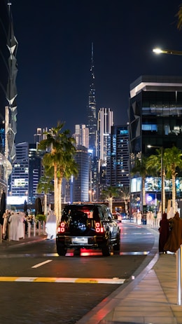 An evening cityscape featuring a vibrant street with a mix of modern skyscrapers, palm trees, and people dressed in traditional attire. A black SUV is traveling down the street, which is lined with illuminated buildings and decorative lights. The scene includes a prominent, illuminated tower in the background.