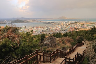 A wooden boardwalk descends through lush greenery towards a coastal town. The town is situated by the sea, with water channels and distant hills visible. Buildings are clustered together, ranging from low-rise to mid-rise structures, giving a charming and colorful appearance. The sky is partly cloudy, casting a serene and calm ambiance.
