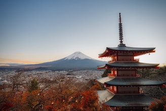 A multilevel pagoda stands in the foreground, surrounded by colorful autumn trees. In the background, Mount Fuji is visible with a snow-capped peak against a clear sky. The cityscape stretches out between the pagoda and the mountain.