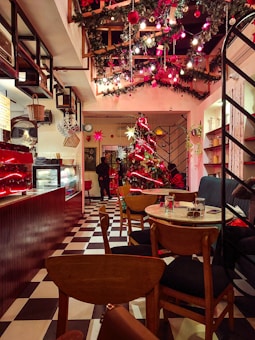 A cozy café decorated for Christmas with a large Christmas tree adorned with red and gold ornaments and lights. The ceiling is adorned with garlands and Christmas lights. There are wooden chairs and tables arranged neatly, and a counter displaying various items. A person stands near the entrance, and the overall ambiance is warm and welcoming.