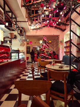 A cozy café decorated for Christmas with a large Christmas tree adorned with red and gold ornaments and lights. The ceiling is adorned with garlands and Christmas lights. There are wooden chairs and tables arranged neatly, and a counter displaying various items. A person stands near the entrance, and the overall ambiance is warm and welcoming.