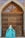 A person wearing a long, flowing turquoise dress with intricate patterns stands in front of a large, ornate wooden door that is part of a building featuring decorative blue and yellow tiles.