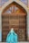 A person wearing a long, flowing turquoise dress with intricate patterns stands in front of a large, ornate wooden door that is part of a building featuring decorative blue and yellow tiles.