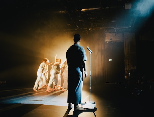 A person stands on a dimly lit stage facing a microphone, while a group of performers in light-colored costumes dance in the background amid theatrical lighting and smoke effects.