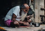 An elderly man with a long gray beard is sitting cross-legged on the ground. He is wearing a white t-shirt and a pink checkered cloth around his waist. He is focused on a smartphone in his hand, and there is a metal pot beside him. The setting appears to be rustic and simple, with wooden walls and various items hanging in the background.