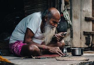 An elderly man with a long gray beard is sitting cross-legged on the ground. He is wearing a white t-shirt and a pink checkered cloth around his waist. He is focused on a smartphone in his hand, and there is a metal pot beside him. The setting appears to be rustic and simple, with wooden walls and various items hanging in the background.