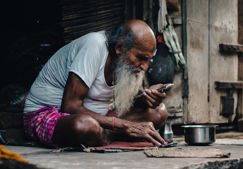 An elderly man with a long gray beard is sitting cross-legged on the ground. He is wearing a white t-shirt and a pink checkered cloth around his waist. He is focused on a smartphone in his hand, and there is a metal pot beside him. The setting appears to be rustic and simple, with wooden walls and various items hanging in the background.
