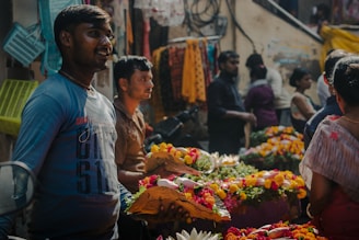 A busy street market scene with vendors selling vibrant flower garlands. People are interacting and moving around in the background, creating a lively atmosphere. Stalls are filled with colorful flowers and other items, and a mix of people are seen engaging with the vendors.