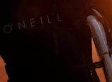 Close-up of a section of a wetsuit with the word 'O'NEILL' visible, and part of a surfboard with an orange light illuminating the scene, suggesting sunset or sunrise.