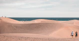 A picturesque desert landscape with vast sand dunes stretching into the distance. The sky is partly cloudy, and the ocean is visible in the background. Several people are scattered across the dunes, some sitting or walking, enjoying the serene setting.