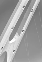 An architectural detail of a bridge featuring large, elongated, curved metal beams intersected by cables set against a clear sky. The image is depicted in grayscale, highlighting the sleek, modern design and smooth surfaces of the structure.