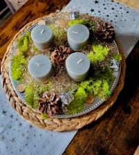 A decorative arrangement featuring four cylindrical candles, likely made of gray wax, placed on a round woven placemat with a silver tray. The candles are surrounded by pine cones, green moss, silver decorative elements, and some wood slices. The setting is on a wooden table with a star-patterned, light-colored fabric underneath.