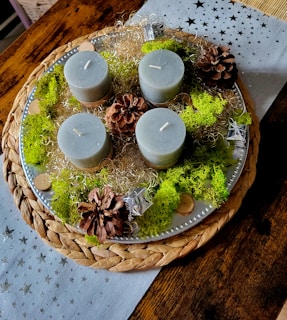 A decorative arrangement featuring four cylindrical candles, likely made of gray wax, placed on a round woven placemat with a silver tray. The candles are surrounded by pine cones, green moss, silver decorative elements, and some wood slices. The setting is on a wooden table with a star-patterned, light-colored fabric underneath.