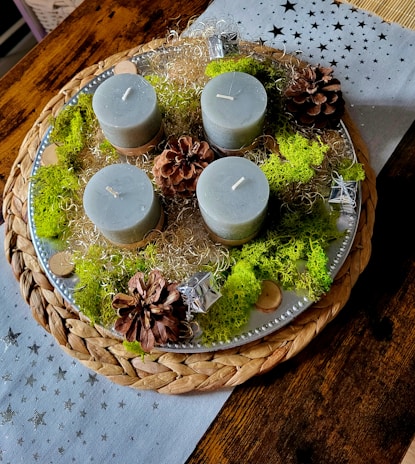 A decorative arrangement featuring four cylindrical candles, likely made of gray wax, placed on a round woven placemat with a silver tray. The candles are surrounded by pine cones, green moss, silver decorative elements, and some wood slices. The setting is on a wooden table with a star-patterned, light-colored fabric underneath.