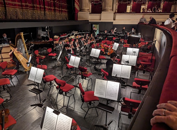 A well-lit orchestra pit with musicians preparing their instruments and sheet music on stands. Red upholstered seats surround the pit, and a grand gilded harp is prominently placed. Musicians are tuning their instruments, while the audience is seen gathering above.