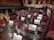 A well-lit orchestra pit with musicians preparing their instruments and sheet music on stands. Red upholstered seats surround the pit, and a grand gilded harp is prominently placed. Musicians are tuning their instruments, while the audience is seen gathering above.