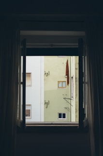 There is an open window framing a view of an exterior building wall. The building has light green and white sections, with small rectangular windows and external fixtures. An orange or brown fabric appears to hang on the wall, possibly part of an awning or cover.