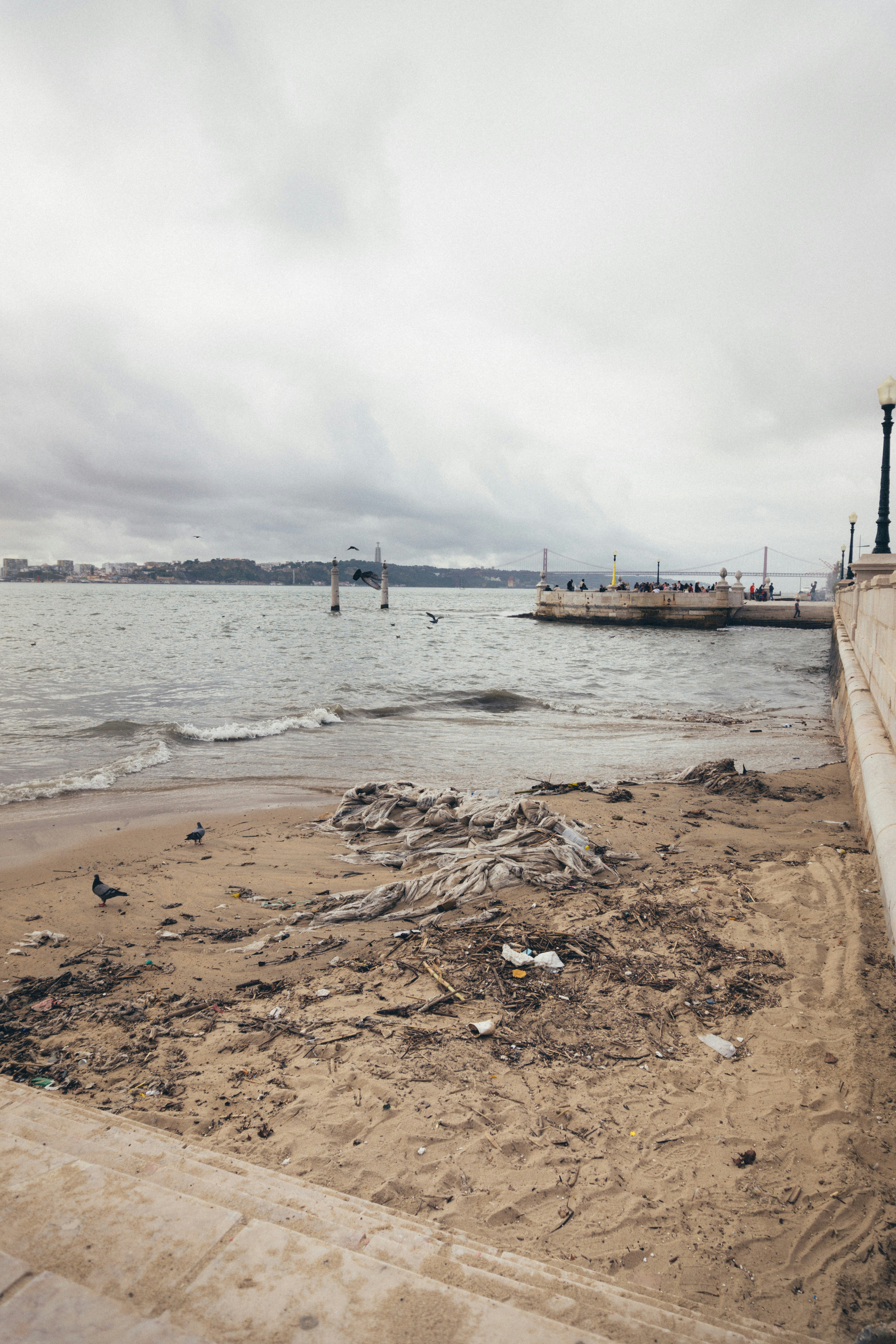 Plastic waste pollution on beach, dark overcast sky, murky grey-green water, debris scattered on sand, dim harsh lighting