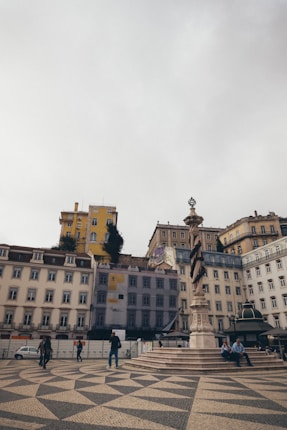 A city square with patterned cobblestones and several people walking or sitting. Surrounding buildings feature a mix of architectural styles, with one yellow building notably standing out. A tall, ornate column stands as a centerpiece in the square.