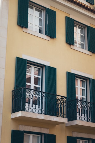 A section of a building facade with three windows featuring dark green shutters and decorative wrought iron balconies. The wall is painted in a warm yellow color, which contrasts with the shutters and balconies.