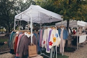 A market stall featuring a collection of colorful winter clothing items displayed on hangers under a white canopy. The garments include jackets and sweaters, some with floral patterns. The scene is set in an outdoor environment surrounded by trees and cobblestone paths, with another stall and a person visible in the background.