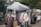 A market stall featuring a collection of colorful winter clothing items displayed on hangers under a white canopy. The garments include jackets and sweaters, some with floral patterns. The scene is set in an outdoor environment surrounded by trees and cobblestone paths, with another stall and a person visible in the background.