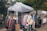 A market stall featuring a collection of colorful winter clothing items displayed on hangers under a white canopy. The garments include jackets and sweaters, some with floral patterns. The scene is set in an outdoor environment surrounded by trees and cobblestone paths, with another stall and a person visible in the background.