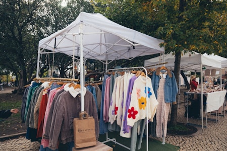 A market stall featuring a collection of colorful winter clothing items displayed on hangers under a white canopy. The garments include jackets and sweaters, some with floral patterns. The scene is set in an outdoor environment surrounded by trees and cobblestone paths, with another stall and a person visible in the background.
